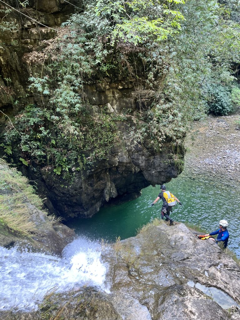 A guest cliff jumps into a deep pool in front of a canyoning guide.