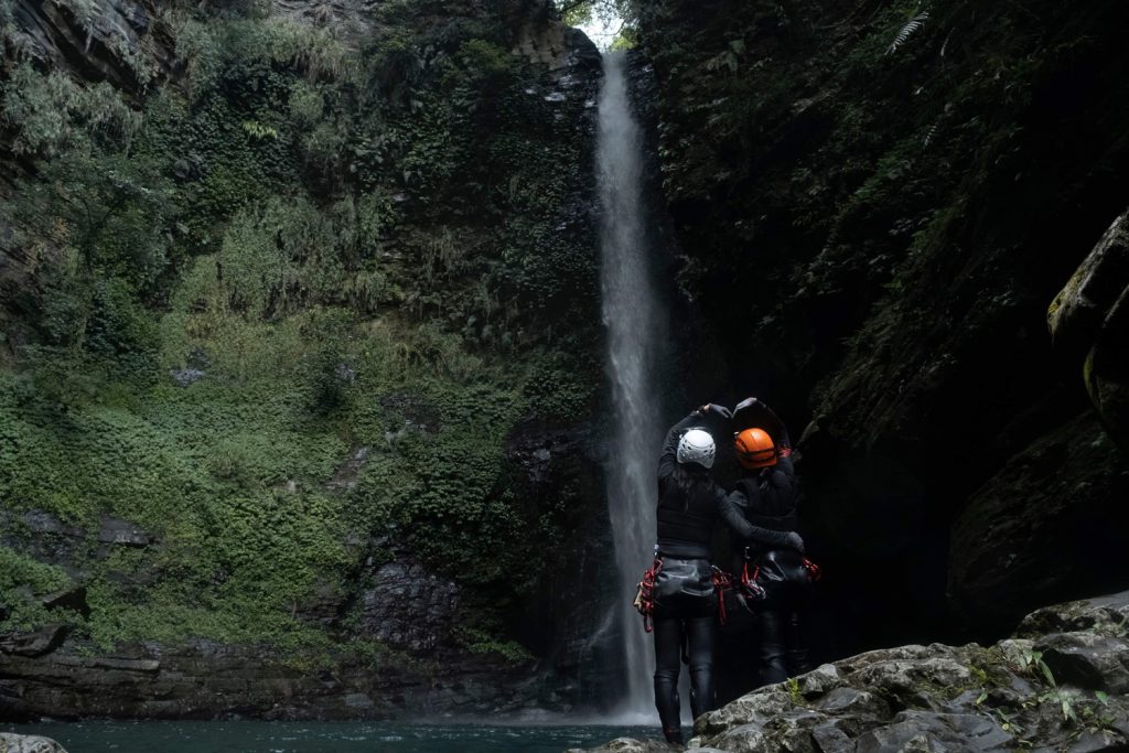 Two people making a heart shape with their hands underneath a tall waterfall.